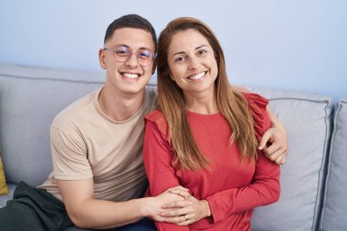 Erkek ve kadın anne ve oğul evdeki kanepede oturup birbirlerine sarılıyorlar.