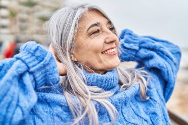 Orta yaşlı, gri saçlı, gülümseyen, kendine güvenen, elleri deniz kenarında rahatlamış bir kadın.