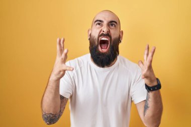 Young hispanic man with beard and tattoos standing over yellow background crazy and mad shouting and yelling with aggressive expression and arms raised. frustration concept. 