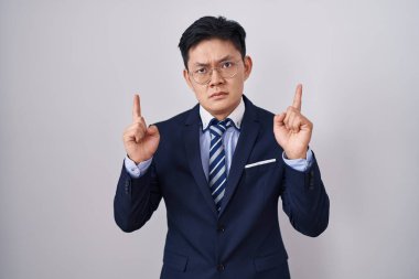 Young asian man wearing business suit and tie pointing up looking sad and upset, indicating direction with fingers, unhappy and depressed. 