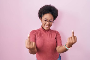 Beautiful african woman with curly hair standing over pink background showing middle finger doing fuck you bad expression, provocation and rude attitude. screaming excited 