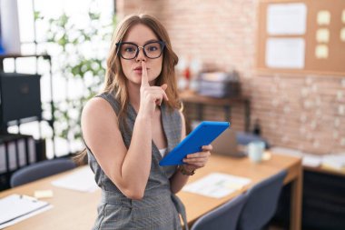 Caucasian woman working at the office wearing glasses asking to be quiet with finger on lips. silence and secret concept. 