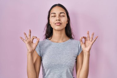 Pembe arka planda rahat bir tişört giyen genç Brezilyalı kadın rahatla ve parmaklarıyla meditasyon hareketi yaparken gözleri kapalı gülümse. yoga konsepti. 