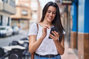 Genç, güzel, İspanyol bir kadın sokakta akıllı telefon kullanarak gülümsüyor.