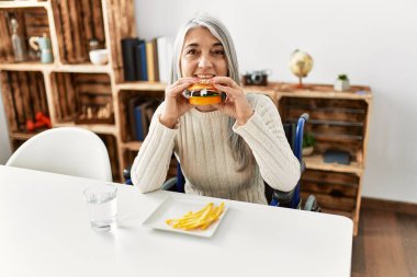 Orta yaşlı, kır saçlı, klasik hamburger yiyen, tekerlekli sandalyede oturan bir kadın.