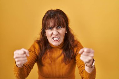 Middle age hispanic woman standing over yellow background angry and mad raising fists frustrated and furious while shouting with anger. rage and aggressive concept. 