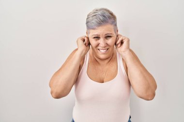 Middle age caucasian woman standing over white background covering ears with fingers with annoyed expression for the noise of loud music. deaf concept. 