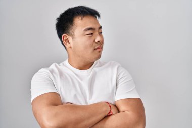 Young chinese man standing over white background looking to the side with arms crossed convinced and confident 