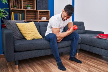 Young hispanic man training using dumbbell sitting on sofa at home