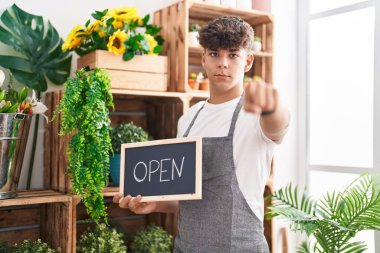 Çiçekçide çalışan İspanyol bir genç elinde açık bir levhayla kamerayı işaret ediyor ve sen kendine güvenen ciddi bir hareket yapıyorsun. 