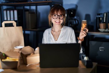 Genç ve güzel bir kadın bilgisayarla çalışıyor ve mutlu ve havalı bir gülümsemeyle yemek yiyor. şanslı kişi. 