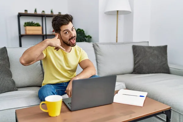 Young man with beard using laptop at home smiling doing phone gesture with hand and fingers like talking on the telephone. communicating concepts.