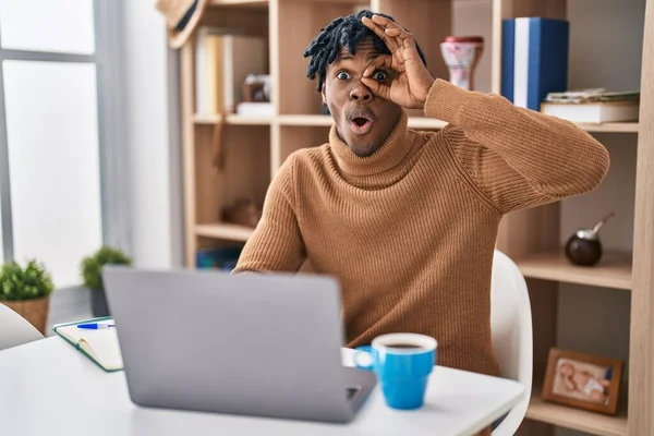 Young african man with dreadlocks working using computer laptop doing ok gesture shocked with surprised face, eye looking through fingers. unbelieving expression.