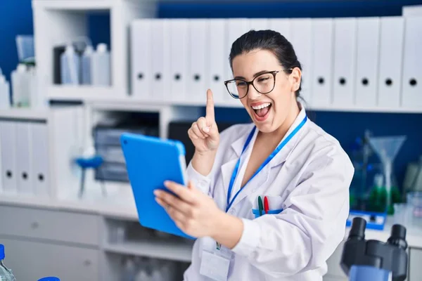Young brunette woman working at scientist laboratory using tablet smiling with an idea or question pointing finger with happy face, number one