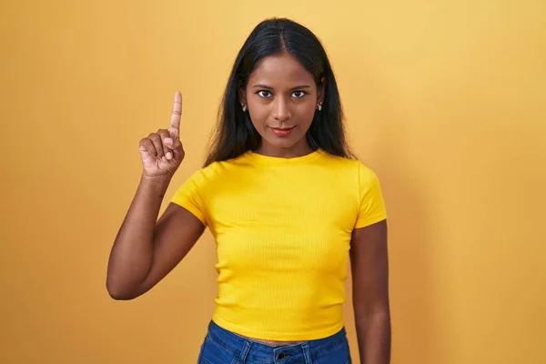 stock image Young indian woman standing over yellow background showing and pointing up with finger number one while smiling confident and happy. 