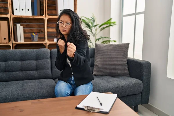 Young asian woman at consultation office ready to fight with fist defense gesture, angry and upset face, afraid of problem