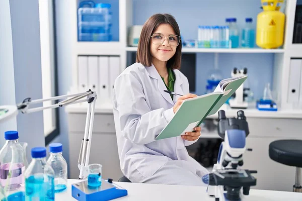Young beautiful hispanic woman scientist smiling confident writing notebook at laboratory