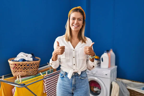 洗濯室の成功サインで若いブロンドの女性は 手で積極的なジェスチャーを行います 親指アップ笑顔と幸せ 陽気な表情と勝者のジェスチャー — ストック写真