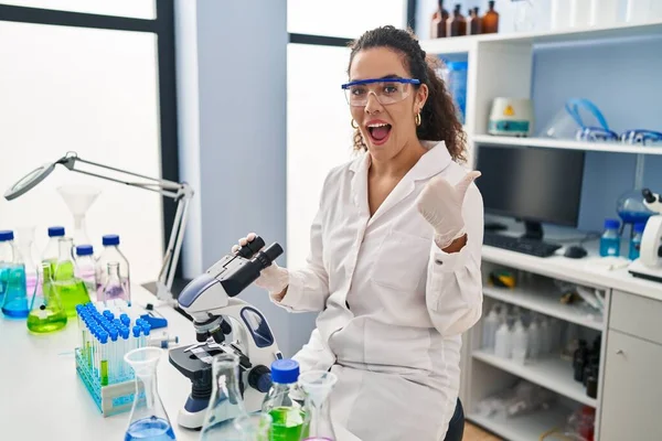 Young hispanic woman working at scientist laboratory pointing thumb up to the side smiling happy with open mouth