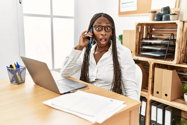 Black woman with braids working at the office speaking on the phone in shock face, looking skeptical and sarcastic, surprised with open mouth
