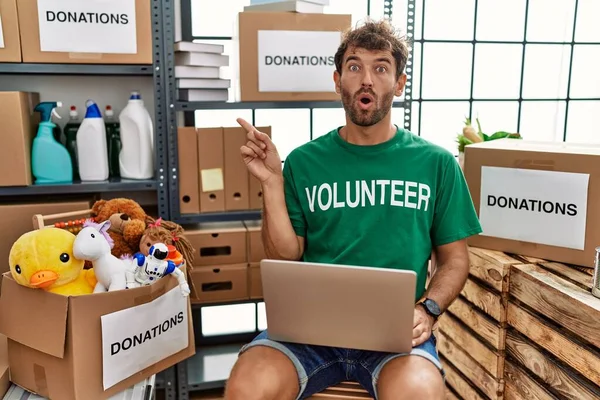 年轻英俊的男子穿着志愿者的T恤 用笔记本电脑惊讶地指指点点旁边 张开嘴惊讶地表情 — 图库照片