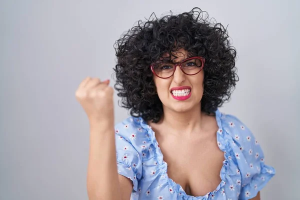 孤立した背景の上に眼鏡をかけている巻き毛の若いブルネットの女性は怒りで叫んでいる間に怒りと怒りに苛まれ 激怒して拳を上げる 怒りと攻撃的な考え方 — ストック写真