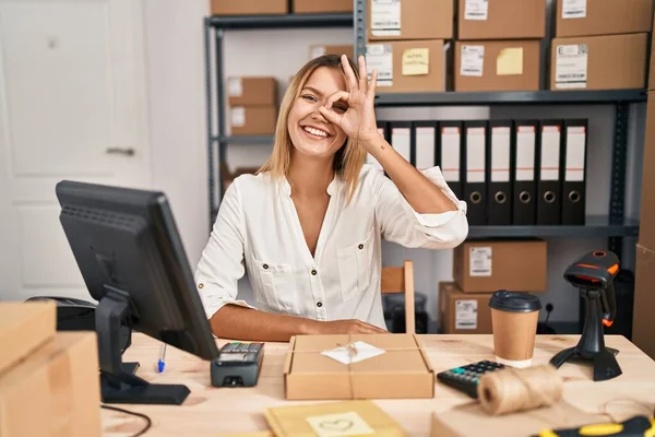 Young blonde woman working at small business ecommerce doing ok gesture with hand smiling, eye looking through fingers with happy face.