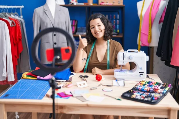 Young hispanic tailor woman doing tutorial with smartphone smiling happy and positive, thumb up doing excellent and approval sign
