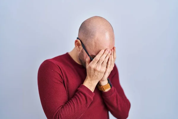 Young bald man with beard standing over white background wearing glasses with sad expression covering face with hands while crying. depression concept.