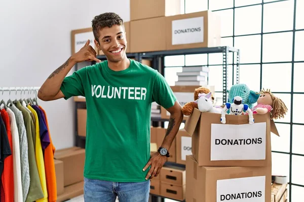 Young handsome hispanic man wearing volunteer t shirt at donations stand smiling doing phone gesture with hand and fingers like talking on the telephone. communicating concepts.