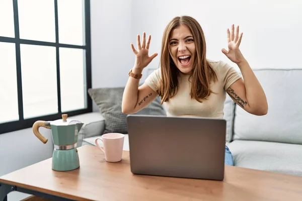 年轻的黑发女人在家里使用笔记本电脑 喝着一杯咖啡庆祝疯狂和惊奇的成功 胳膊抬起 睁开眼睛尖叫兴奋 获奖者概念 — 图库照片