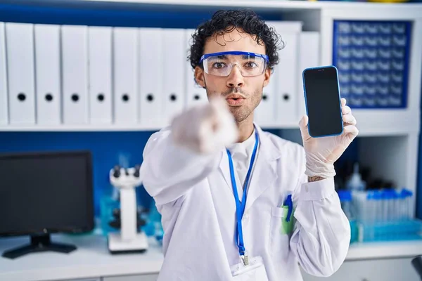 Hispanic man working at scientist laboratory showing smartphone screen pointing with finger to the camera and to you, confident gesture looking serious