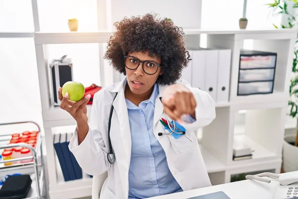 Black doctor woman with curly hair holding green apple pointing with finger to the camera and to you, confident gesture looking serious