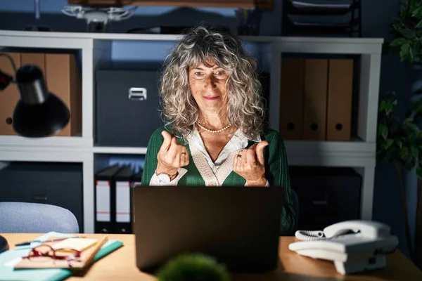 Middle age woman working at night using computer laptop doing money gesture with hands, asking for salary payment, millionaire business