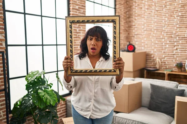 Hispanic woman at new home holding empty frame in shock face, looking skeptical and sarcastic, surprised with open mouth 