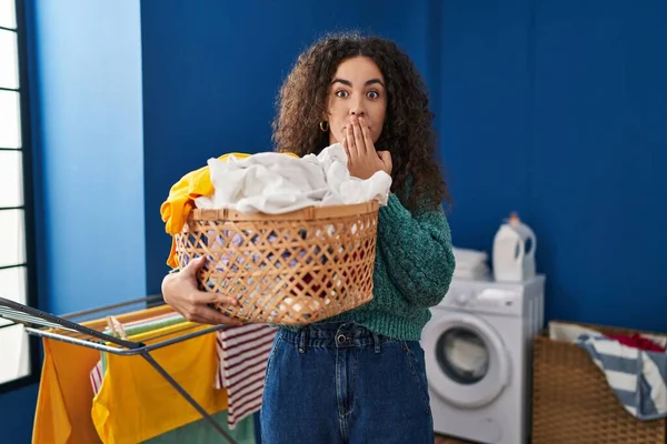 Young Hispanic Woman Holding Laundry Basket Covering Mouth Hand Shocked — Stockfoto