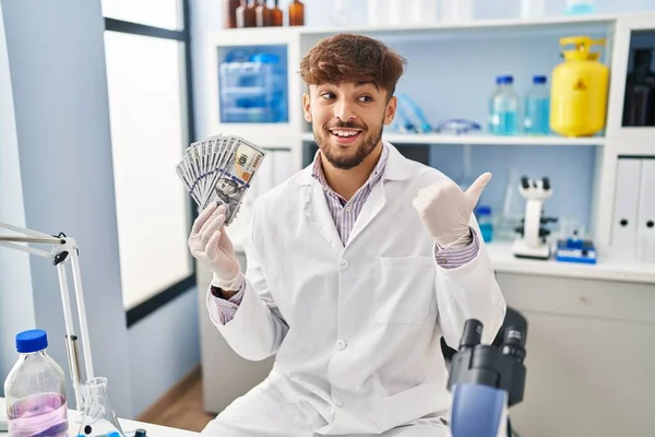 Arab man with beard working at scientist laboratory holding money pointing thumb up to the side smiling happy with open mouth