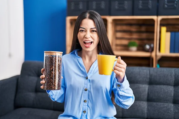 Young brunette woman holding coffee beans and cup of coffee angry and mad screaming frustrated and furious, shouting with anger. rage and aggressive concept. 