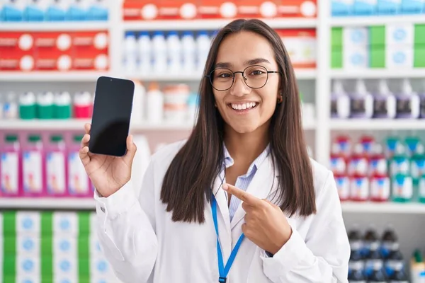 Young hispanic woman working at pharmacy drugstore showing smartphone screen smiling happy pointing with hand and finger