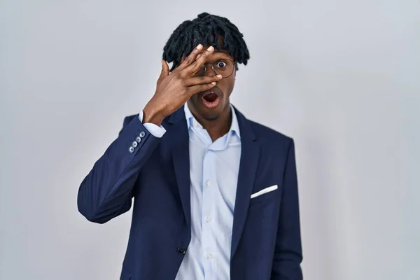 Young african man with dreadlocks wearing business jacket over white background peeking in shock covering face and eyes with hand, looking through fingers with embarrassed expression.