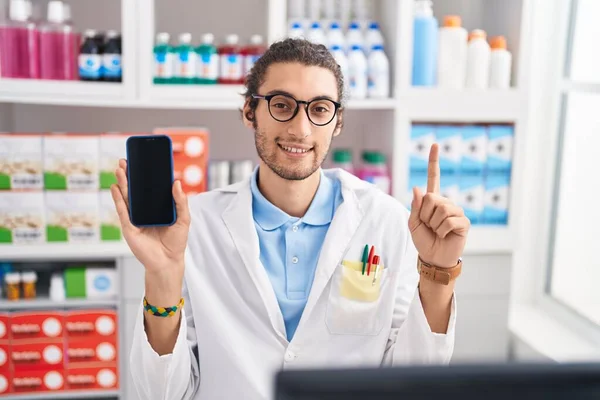 Young hispanic man working at pharmacy drugstore showing smartphone screen smiling with an idea or question pointing finger with happy face, number one