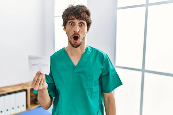 Young hispanic doctor man holding covid record card in shock face, looking skeptical and sarcastic, surprised with open mouth