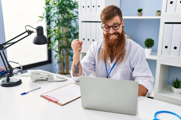 Redhead man with long beard wearing doctor uniform working using computer laptop screaming proud, celebrating victory and success very excited with raised arm