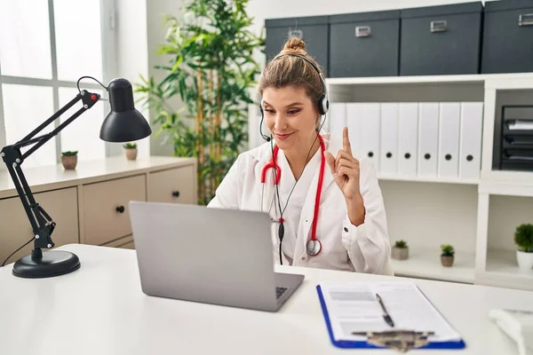 Young doctor woman wearing doctor uniform working using computer laptop surprised with an idea or question pointing finger with happy face, number one