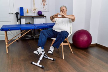 Fizyoterapi kliniğinde aşkta gülümseyen ve elleriyle kalp sembolü yapan pedal egzersizi kullanan kıdemli beyaz adam. romantik kavram. 