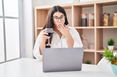 Genç esmer kadın laptopuyla video görüşmesi yapıyor. Ağzı elle kaplayan bir bardak şarap içiyor. Şok olmuş ve hata yapmaktan korkuyor. şaşırmış ifade 