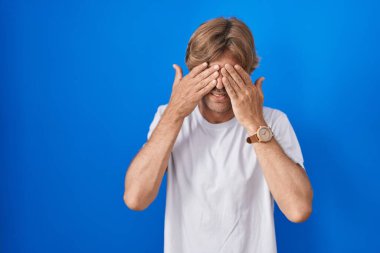 Orta yaşlı bir adam mavi arka planda duruyor. Yorgunluk ve baş ağrısı için gözlerini ovuşturuyor. Uykulu ve yorgun bir ifade. görme sorunu 