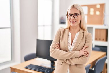 Sarışın, genç bir iş kadını, kollarını kavuşturup ofiste el kol hareketi yapıyor.