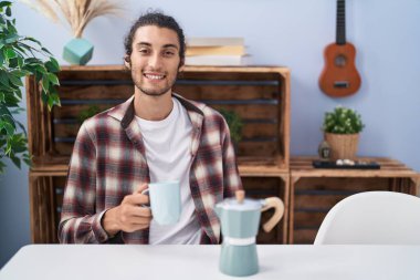 Young hispanic man drinking coffee from french coffee maker looking positive and happy standing and smiling with a confident smile showing teeth 