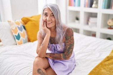 Orta yaşlı, kır saçlı, gülümseyen, yatakta kendine güvenen bir kadın.